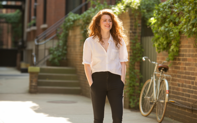 A young woman with curly red hair stands in a sunlit alley, wearing a white shirt and black pants, hands in pockets.